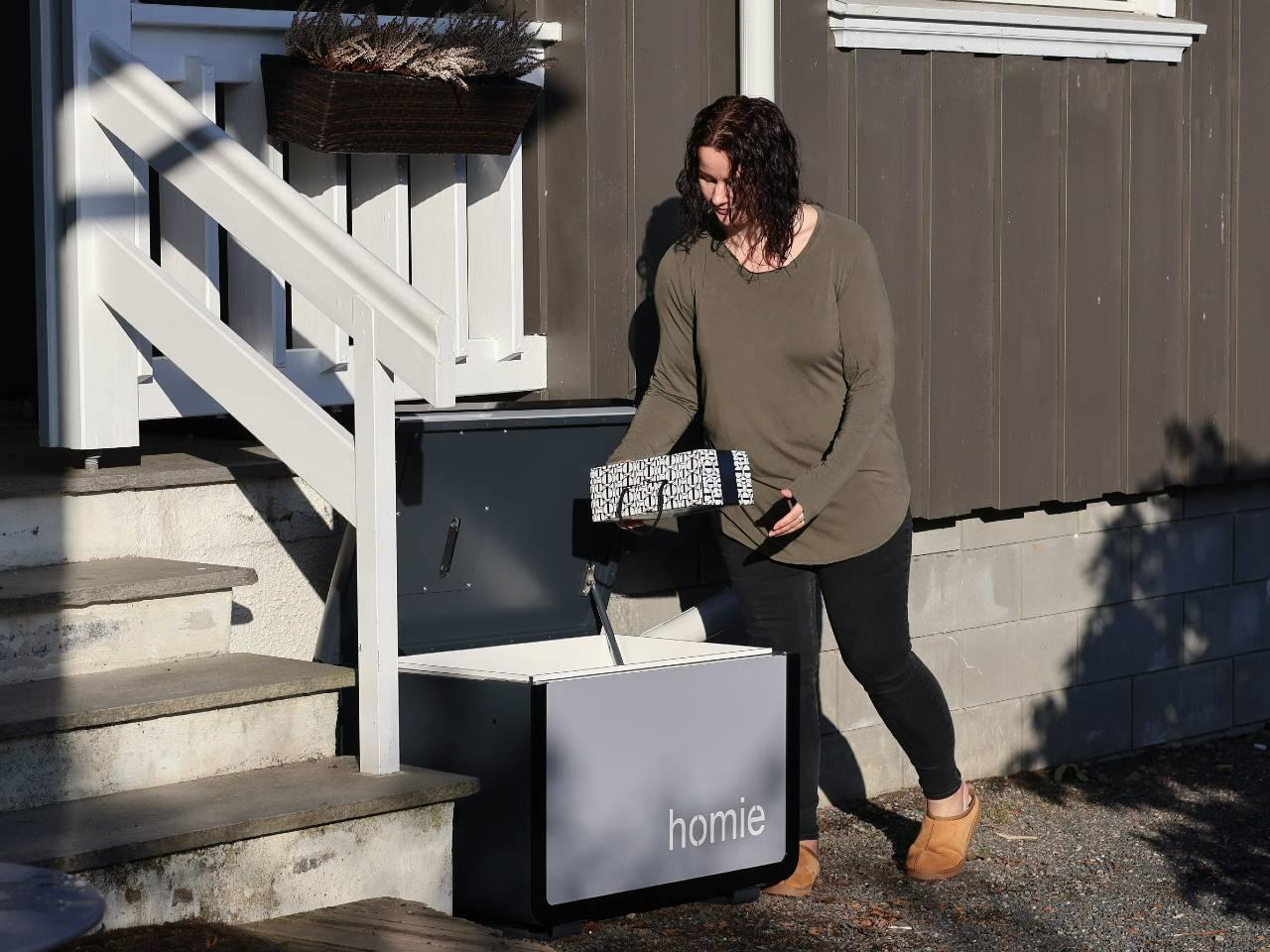 Homie hjemleveringskasse for pakker og matkasser, en kvinne som åpner og tar ut en pakke ved trappen foran huset FOTO