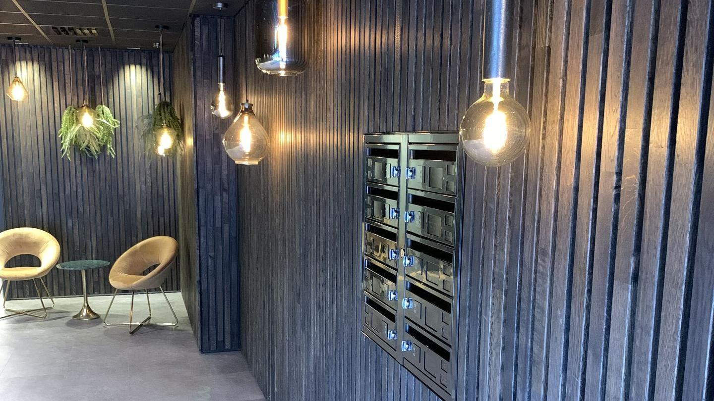 Sort postkassesystem innfelt i trespilevegg, hengende taklys og stoler i et fellesområde FOTO